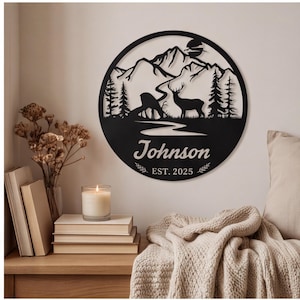 May include: A circular black metal wall sign featuring a laser-cut mountain landscape with pine trees, a winding river, and two deer silhouettes. The text "Johnson" is centered below the scene, followed by the text "EST. 2025" with leaf accents.