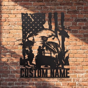 May include: Black metal wall art featuring an American flag, an eagle, and silhouettes of soldiers and a vehicle. The words "CUSTOM NAME" are at the bottom. The art is mounted on a brick wall.