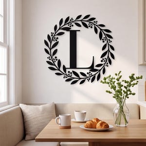 May include: A black metal wall sign featuring a large capital letter L centred within a circular wreath of leaves and small berries. The design is flat and symmetrical, showcasing a botanical motif that surrounds the central character.
