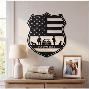 Può includere: Una targa da parete in metallo nero a forma di scudo con un motivo ritagliato della bandiera americana. Il design include le sagome di due figure, un'auto della polizia e due cani, con una barra orizzontale contenente il testo CUSTOM TEXT.