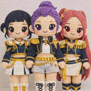May include: Three crocheted dolls in navy and gold uniforms, each with a unique hairstyle: dark blue, purple, and red. The dolls have smiling faces and wear white skirts or shorts. The dolls are handmade.