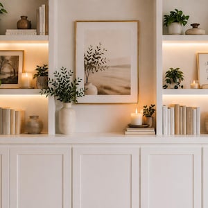 Könnte beinhalten: Ein weißer Einbauschrank mit offenen Regalen, der gerahmte Kunstwerke, Bücher, Kerzen und Topfpflanzen zeigt. Die Regale sind mit warmer Beleuchtung versehen, was eine gemütliche und einladende Atmosphäre schafft.