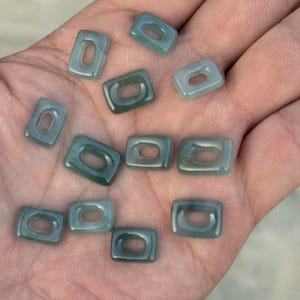 Puede incluir: Una colección de pequeñas cuentas rectangulares de color verde azulado con un agujero rectangular redondeado en el centro. Las cuentas son lisas y brillantes, y se sostienen en una mano abierta.