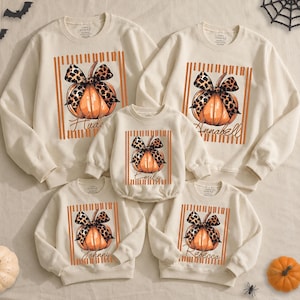 May include: A set of five cream-colored long-sleeve sweatshirts and a baby bodysuit featuring a graphic of an orange pumpkin with a leopard print bow. Each garment displays a vertical orange striped square background and a unique name in script.