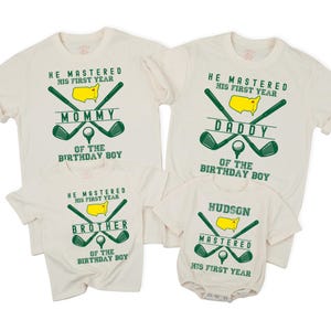 May include: Off-white family golf-themed t-shirts and a onesie. Each shirt features a green golf club design, a yellow map, and text that reads "Mommy", "Daddy", "Brother", and "Hudson". The shirts also say "He mastered his first year".