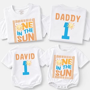 May include: Four white garments including two short-sleeve t-shirts and two baby bodysuits. The designs feature orange and blue text reading "ONE IN THE SUN" or "DADDY 1" or "DAVID 1" with a small smiling sun graphic on each piece of clothing.