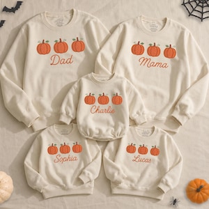 May include: Five cream-colored long-sleeve sweatshirts featuring embroidered orange pumpkins and personalized names. The shirts display the text Dad, Mama, Charlie, Sophia, and Lucas in cursive script, with three pumpkins above each name.