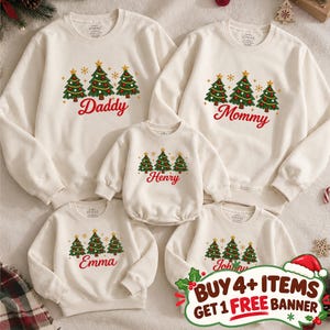 May include: A set of five off-white long-sleeve sweatshirts featuring a graphic of three green Christmas trees with gold stars and red ornaments. Each sweatshirt displays a unique name in red script: Daddy, Mommy, Henry, Emma, and Johnny.