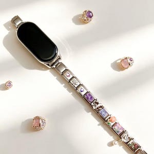 Peut inclure: Un bracelet de montre intelligente argenté orné de breloques décoratives avec des pierres précieuses et des motifs floraux. L'image présente également plusieurs bagues en pierres précieuses roses, violettes et dorées.