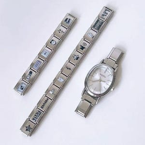 Puede incluir: Tres pulseras plateadas y un reloj sobre una superficie blanca. Las pulseras están hechas de eslabones rectangulares con varios iconos pequeños. El reloj tiene una correa plateada y una esfera ovalada blanca con la palabra "NAIDU".
