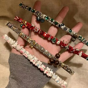 May include: Three link bracelets featuring distinct designs. One bracelet has white bear charms, another is red with holiday-themed charms like trees and bells, and the third is silver-toned with green and black gemstone-style accents and charms.