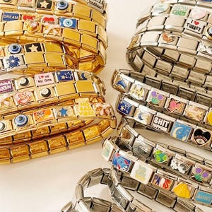 Puede incluir: Montones de pulseras de abalorios doradas y plateadas con varios abalorios decorativos. Los abalorios presentan diseños como estrellas, corazones y frases como "DREAM" y "NO BAD VIBES". Algunos abalorios tienen detalles de esmalte de colores.