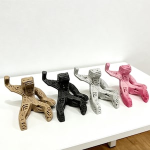 May include: Four abstract human-like sculptures in a row. The sculptures are in shades of tan, black, white, and pink. Each figure has a geometric pattern carved into its surface, with one arm raised.