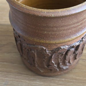 Puede incluir: Un vaso cilíndrico de cerámica con un interior liso de color amarillo mostaza y un exterior texturizado de color marrón oscuro. La parte inferior del recipiente muestra un patrón de relieve orgánico, rugoso y elevado que contrasta con el borde superior liso.