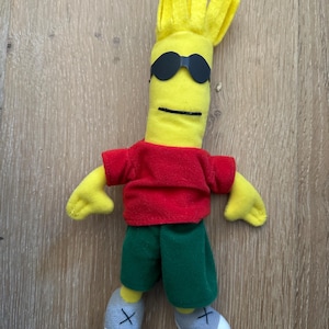 May include: A plush doll with yellow skin, spiky yellow hair, and black sunglasses. The doll wears a red short-sleeved shirt, green shorts, and grey shoes with black X-shaped stitching on the front. The facial features include a simple black line mouth.