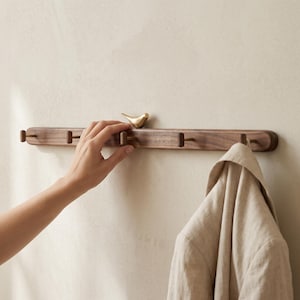 May include: A wooden wall-mounted coat rack with four pegs and a small brass bird figurine. The rack is made of dark wood and has a curved design. A beige jacket hangs from one of the pegs.