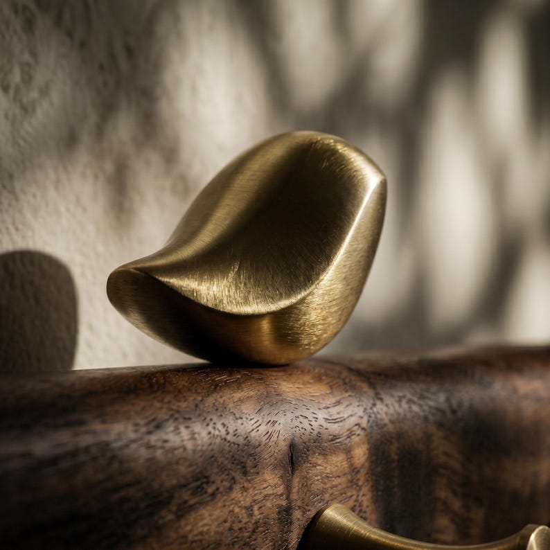 May include: A brushed brass bird-shaped decorative object with a smooth, metallic finish. The bird figure is perched atop a dark, textured wooden surface that features visible wood grain patterns and a warm, natural brown tone.