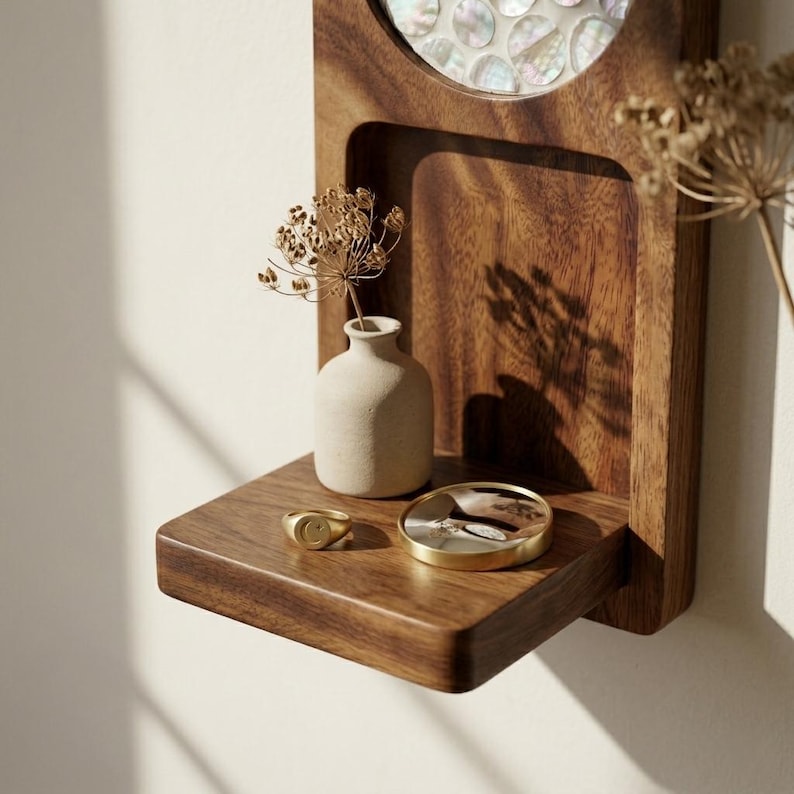 May include: A wooden wall shelf with a small, beige vase holding dried flowers. A gold ring and a small, round, gold-rimmed dish sit on the shelf. The shelf is made of dark wood and has a built-in mirror.