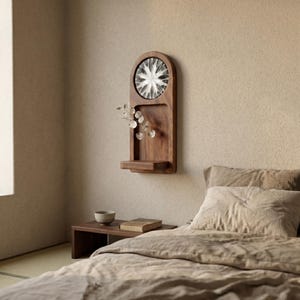 May include: A wooden wall-mounted shelf with an arched top, featuring a circular, starburst-like design. The shelf includes a small ledge and holds delicate, clear decorative stems. The bedroom setting includes a bed, side table, and a window.