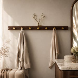 Peut inclure: Un porte-manteau mural en bois foncé avec six boutons en laiton. Une robe et une serviette beiges sont suspendues aux crochets. Un porte-bijoux décoratif en laiton en forme d'arbre est au-dessus. La salle de bain comprend un comptoir en marbre et un miroir.