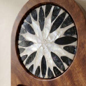 May include: A wooden decorative piece with a circular inlay. The inlay features a floral design made of black and white shell pieces. The wooden frame is a warm brown color, and the shell pieces have a pearlescent sheen.