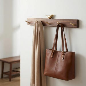 May include: A wooden wall-mounted coat rack with four brass hooks and a decorative brass bird. A tan scarf and a brown leather tote bag hang from the hooks. The rack is mounted on a white wall.