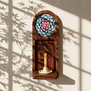 May include: A wooden wall-mounted candle holder with a decorative mosaic tile design in shades of blue, purple, and white. The holder features a small shelf with a lit candle in a cream-colored holder. The wood has a rich, dark brown finish.