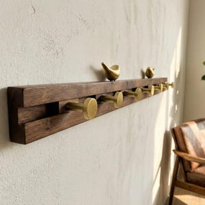 May include: A wooden wall-mounted coat rack with six brass hooks. Two small, gold-colored bird sculptures sit on the top of the rack. The wood is a dark brown color, and the hooks are cylindrical with a flat, round front.