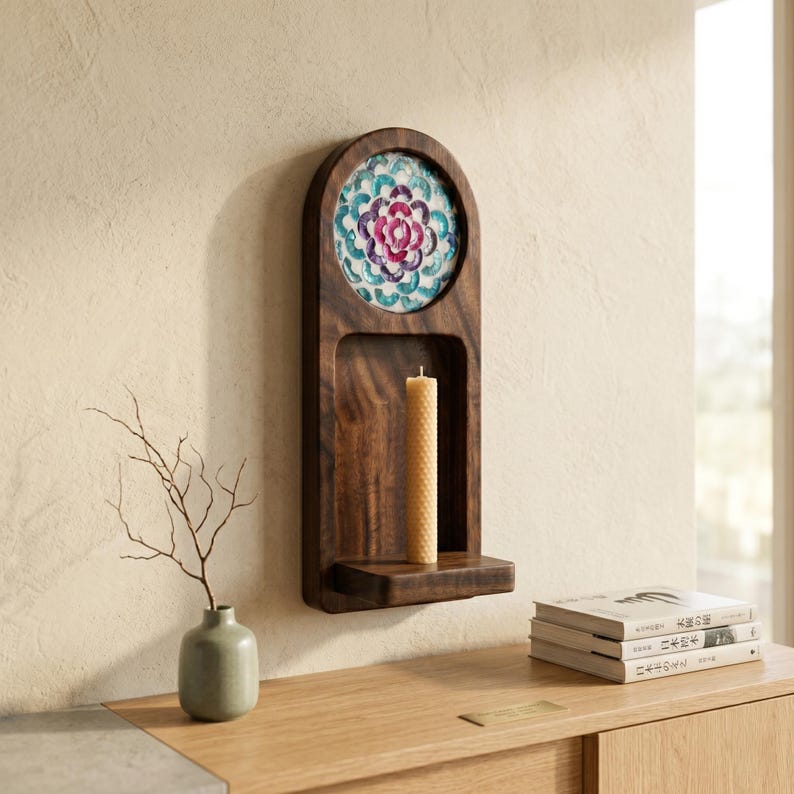 May include: A dark wood wall sconce with a floral design and a shelf. The sconce has a circular design with a colorful floral pattern in the center. A tall, cylindrical candle sits on the shelf. A small vase with a branch is on a wooden table.