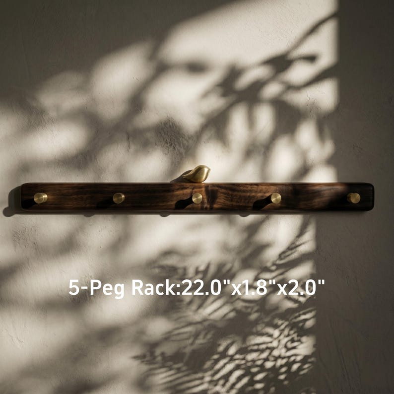 May include: A dark wood wall-mounted rack featuring five brass pegs and a small brass bird ornament perched on top. The rack measures 22.0 inches by 1.8 inches by 2.0 inches, as indicated by the text 5-Peg Rack: 22.0"x1.8"x2.0".
