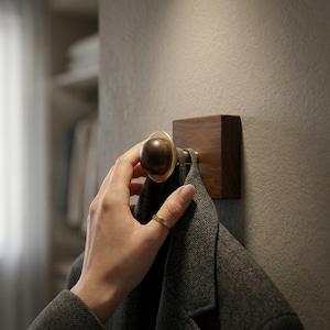 Könnte beinhalten: Ein hölzerner Wandhaken mit Messingakzent und einem dunkelbraunen Knauf. Der Haken ist an einem quadratischen Holzblock befestigt. Ein grauer Mantel hängt an dem Haken.