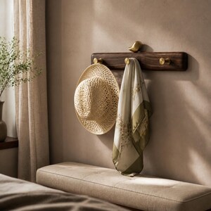 May include: A dark wood wall-mounted coat rack featuring three cylindrical brass hooks and a small brass bird ornament on top. A woven straw hat and a light-colored scarf with a green floral pattern are hanging from the brass hooks.