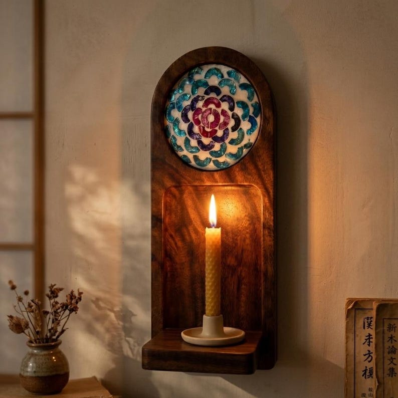 May include: A wooden wall sconce with a decorative mosaic top in blue, teal, and purple. A lit candle sits in the center, casting a warm glow. The sconce has a shelf at the bottom and is mounted on a light-colored wall.