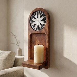 May include: A wooden wall sconce with a candle and decorative inlay. The sconce is made of dark wood and features a circular inlay with a black and white floral design. A cream-colored candle sits on a small shelf.