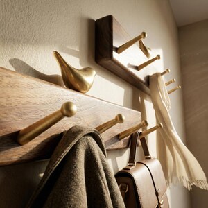 May include: Wooden wall-mounted coat rack with brass hooks and a decorative brass bird. A brown coat, a tan scarf, and a brown leather bag hang from the hooks. The rack is made of dark wood.