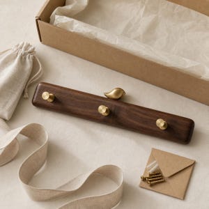 May include: A rectangular dark brown wooden wall hook rack featuring three small round brass knobs and a single brass bird-shaped decorative element on top. The set includes a small brown envelope containing metal screws and wall anchors.