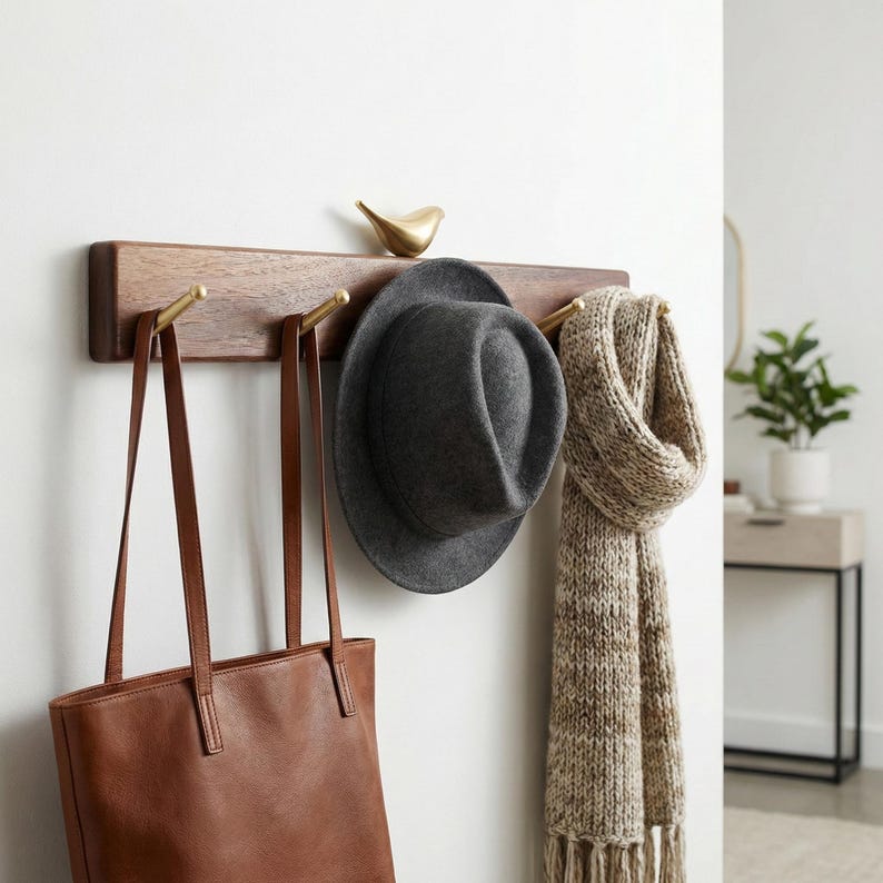 May include: A wooden wall-mounted coat rack with brass hooks and a decorative bird. A gray hat, a brown leather tote bag, and a beige scarf hang from the hooks. The rack is mounted on a white wall.