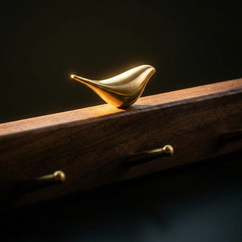 May include: A golden bird-shaped decorative element perched on a dark wooden surface. The bird has a smooth, polished finish and is positioned above three small, round, golden knobs. The wood has a rich, warm tone, and the background is dark.