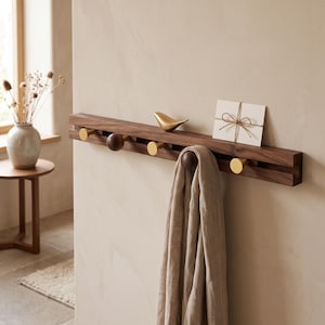 May include: A horizontal wall-mounted coat rack made of dark brown wood featuring a central groove. It includes alternating round brass and dark wood pegs. A small brass bird figurine and a white envelope tied with twine sit on the top edge.