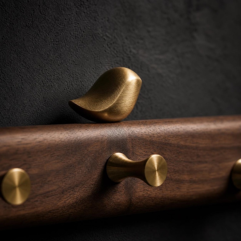 May include: A wall-mounted rack made of dark brown wood featuring a smooth, brushed brass bird-shaped decorative element and several cylindrical brass hooks. The hardware has a matte finish and is attached to a horizontal wooden base.