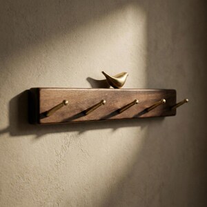 May include: A wooden wall-mounted coat rack with four brass hooks. A small, gold-colored bird figurine sits on the top shelf. The rack is made of dark brown wood and is mounted on a light brown wall.