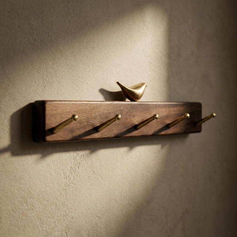 May include: A wooden wall-mounted coat rack with four brass hooks. A small, gold-colored bird figurine sits on the top shelf. The rack is made of dark brown wood and is mounted on a light brown wall.