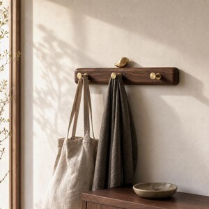 May include: A horizontal dark wood wall-mounted coat rack featuring three circular brass hooks and a small brass bird figurine perched on the top center edge. A beige canvas tote bag and a dark gray fabric cloth hang from the brass hooks.