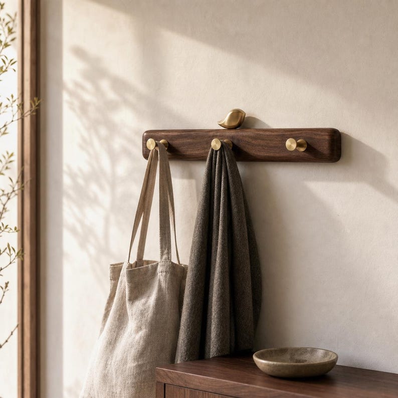 May include: A horizontal dark wood wall-mounted coat rack featuring three circular brass hooks and a small brass bird figurine perched on the top center edge. A beige canvas tote bag and a dark gray fabric cloth hang from the brass hooks.