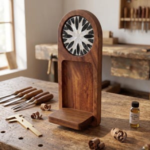 May include: A handcrafted wooden display stand with a decorative inlaid design. The stand features a circular inlay with a starburst pattern. The stand is made of dark wood and sits on a wooden workbench with tools and a bottle of walnut oil.