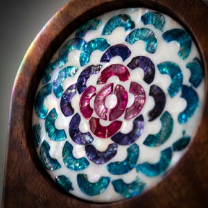May include: Close-up of a decorative wooden object with a central design of iridescent, shell-like pieces in shades of teal, purple, and pink. The design is set against a white background and framed by a dark wood border.