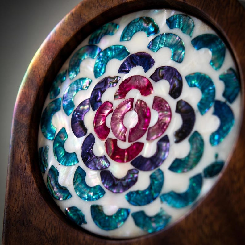May include: Close-up of a decorative wooden object with a central design of iridescent, shell-like pieces in shades of teal, purple, and pink. The design is set against a white background and framed by a dark wood border.