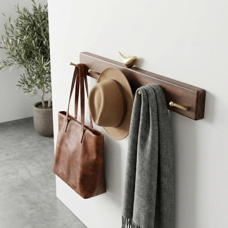 May include: A dark brown wooden wall-mounted coat rack with brass hooks and a small gold bird sculpture. A brown leather tote bag, a tan fedora hat, and a gray scarf are hanging from the rack, creating a stylish storage solution.