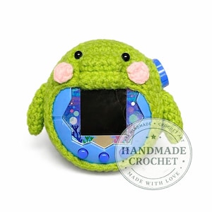 May include: A green crocheted cover shaped like a cartoon character, with pink cheeks, covering a blue and black electronic device. The device has a screen and buttons. A circular sticker reads "HANDMADE CROCHET MADE WITH LOVE".