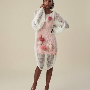 Puede incluir: Vestido blanco de punto calado con mangas largas y voluminosas. El vestido presenta un vestido interior rosa y está adornado con pompones rosas y blancos. La modelo lleva tacones rosas a juego.